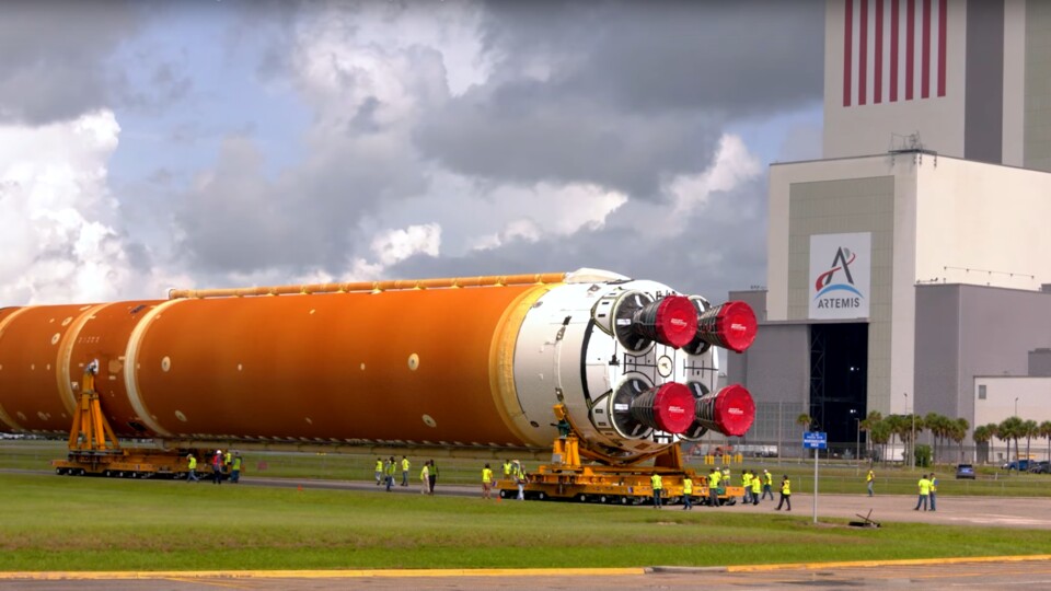 Eine Erststufe des SLS wird angeliefert. Sein Ziel ist das sich gerade öffnende Seitentor – ein Zwerg gegen seine vier Kumpanen auf der Vorder- sowie Rückseite des VABs. (Bildquelle: NASA)